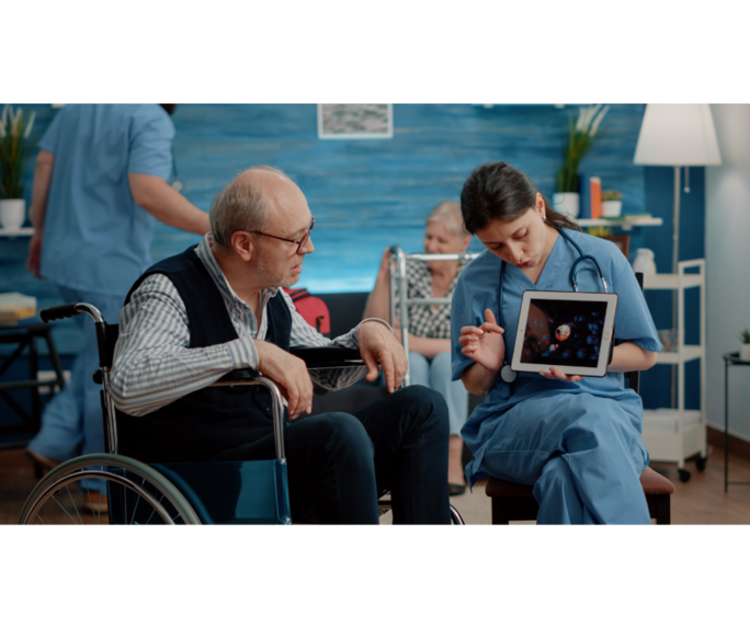 Eine Pflegerin zeigt einem älteren Mann, der im Rollstuhl sitzt, etwas auf einem Tablet.