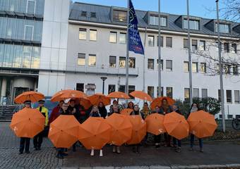 Foto mit Gruppe von Personen im Freien; alle haben orangefarbene Schirme aufgespannt, im Hintergrund eine blaue Flagge mit der Aufschrift: "Frei leben - ohne Gewalt"; im Hintergrund das Landratsamt Stendal © gemeinsam gegen Gewalt