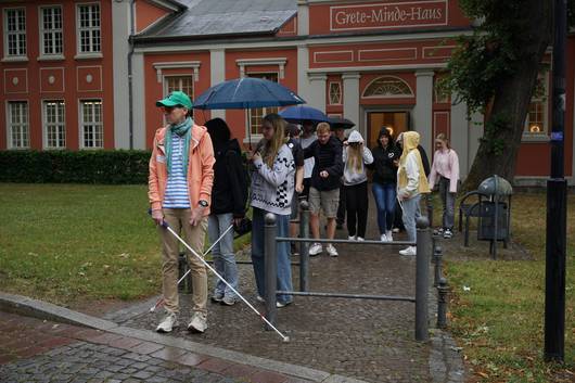 Daniela Waiß vom Blinden- und Sehbehindertenverband steht mit den Schülerinnen und Schülern vor dem Grete-Minde-Saal und möchte mit Langstock die Straße überqueren.